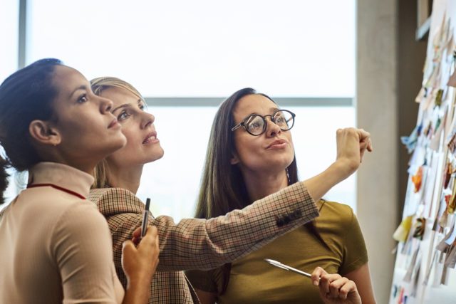 Trois femmes regardent un tableau rempli de post-it