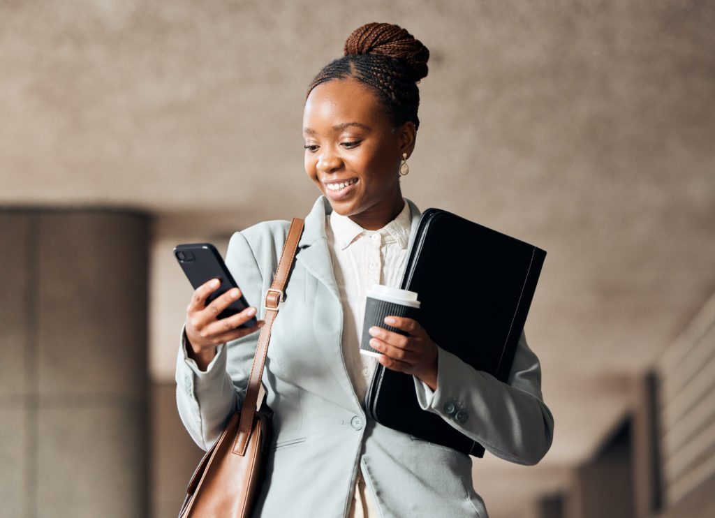 Femme active noire, qui regarde en souriant son téléphone qu'elle tient dans sa main droite, et porte contre elle de son bras gauche une ordinateur, un café à la main